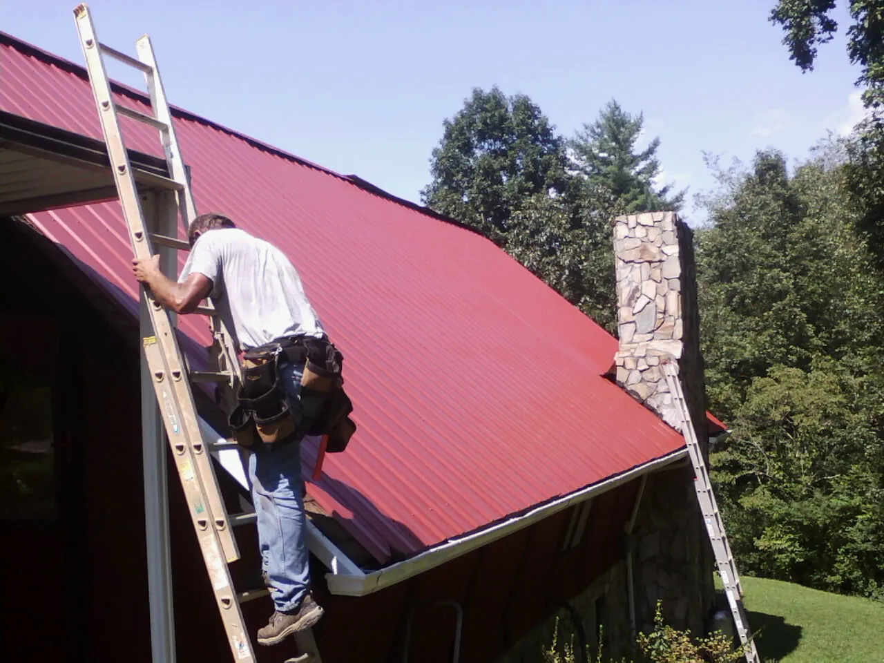 Roofer on a ladder beside a red metal roof and stone chimney — Soffit & Fascia Repair by Roofing Brothers in Tennessee Colony